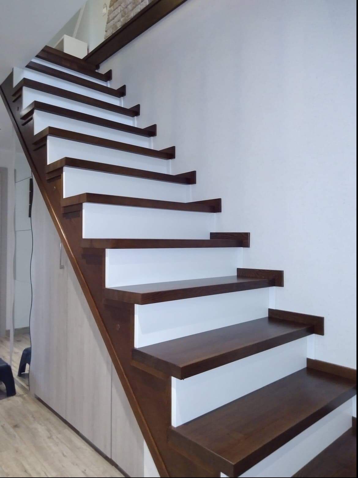 Nowoczesne schody drewniane z ciemnego drewna i białymi podstopnicami, zabudowane szafą z jasnego drewna, widok z dołu.