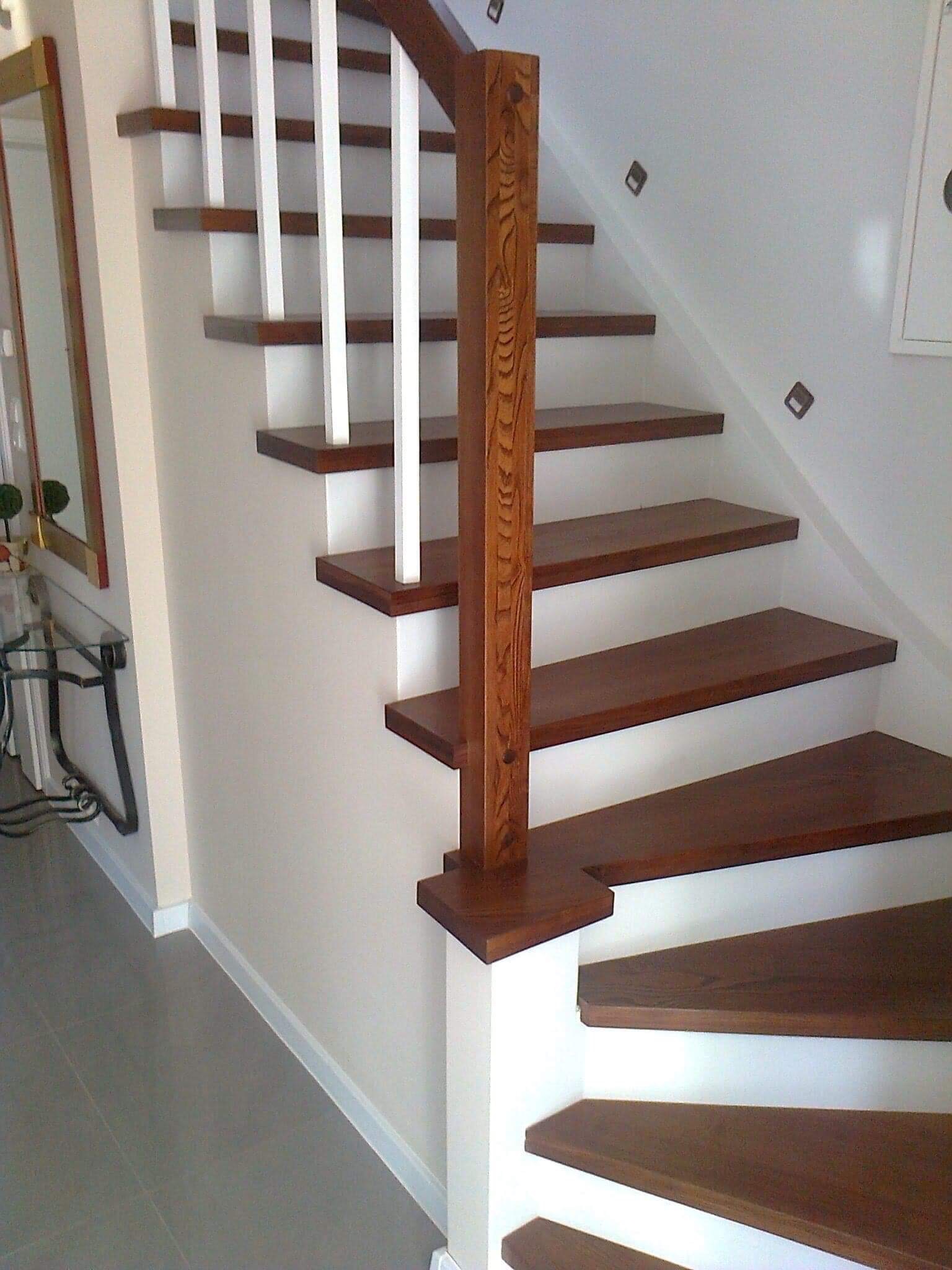 Drewniane schody z białymi balustradami, widok z dołu na ciemnobrązowe stopnie i ozdobny słupek poręczy, na ścianie dekoracyjne kwadratowe naklejki.