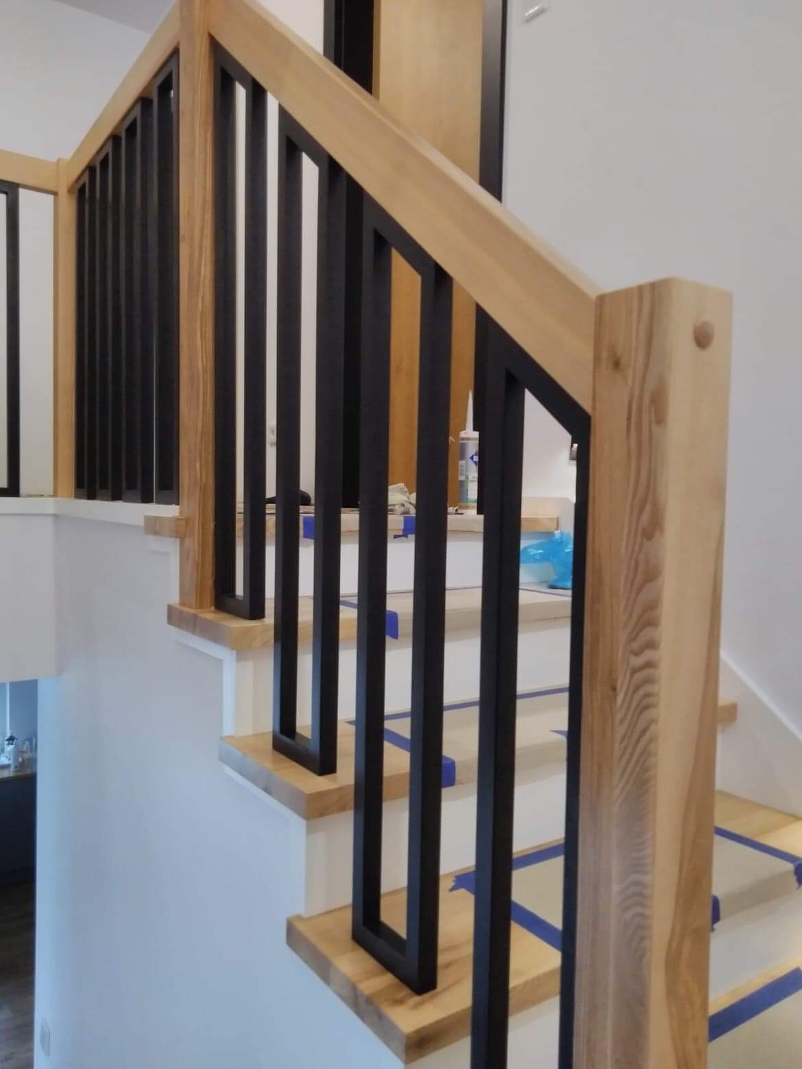 Drewniane schody z czarną, metalową balustradą w trakcie montażu, zabezpieczone taśmą malarską.