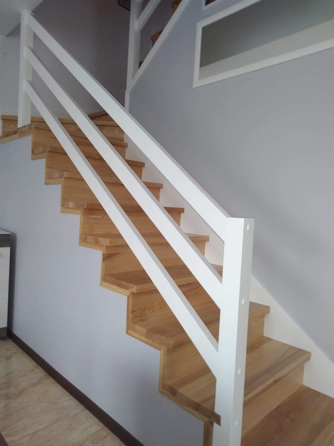 Drewniane schody z białymi, prostymi barierkami w nowoczesnym domu, widoczne trzy stopnie i fragment ściany z obrazem.