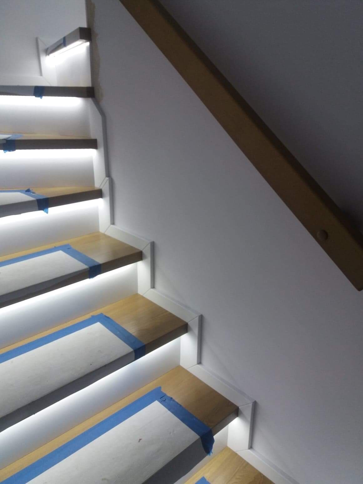 Schody drewniane z podświetleniem LED na stopniach, zabezpieczone taśmą malarską i folią ochronną podczas remontu, widoczna drewniana poręcz.