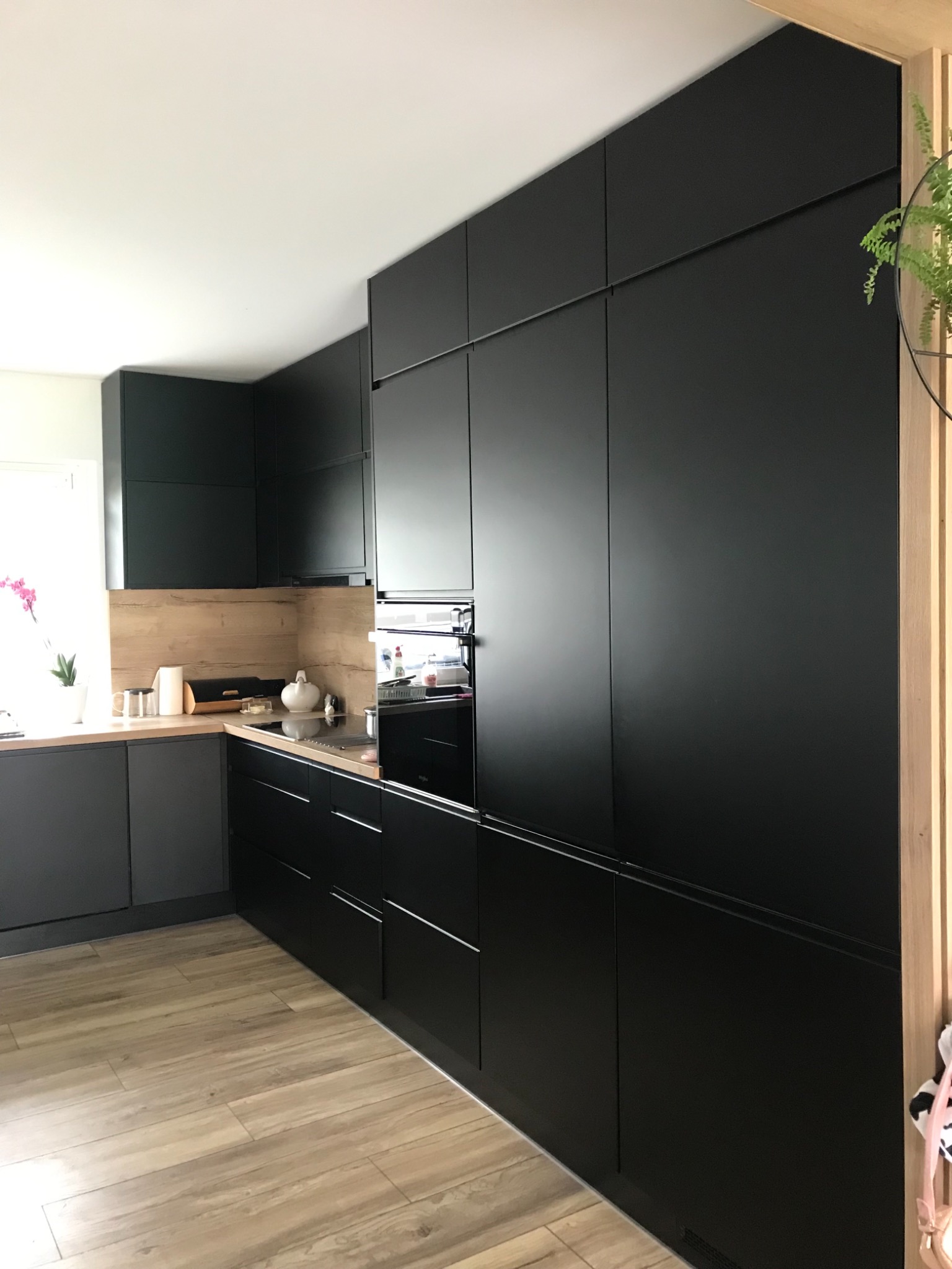 Nowoczesna kuchnia z zabudową w matowej czerni, drewnianymi blatami i panelami ściennymi, uzupełniona o drewniane akcenty dekoracyjne i roślinę wiszącą.