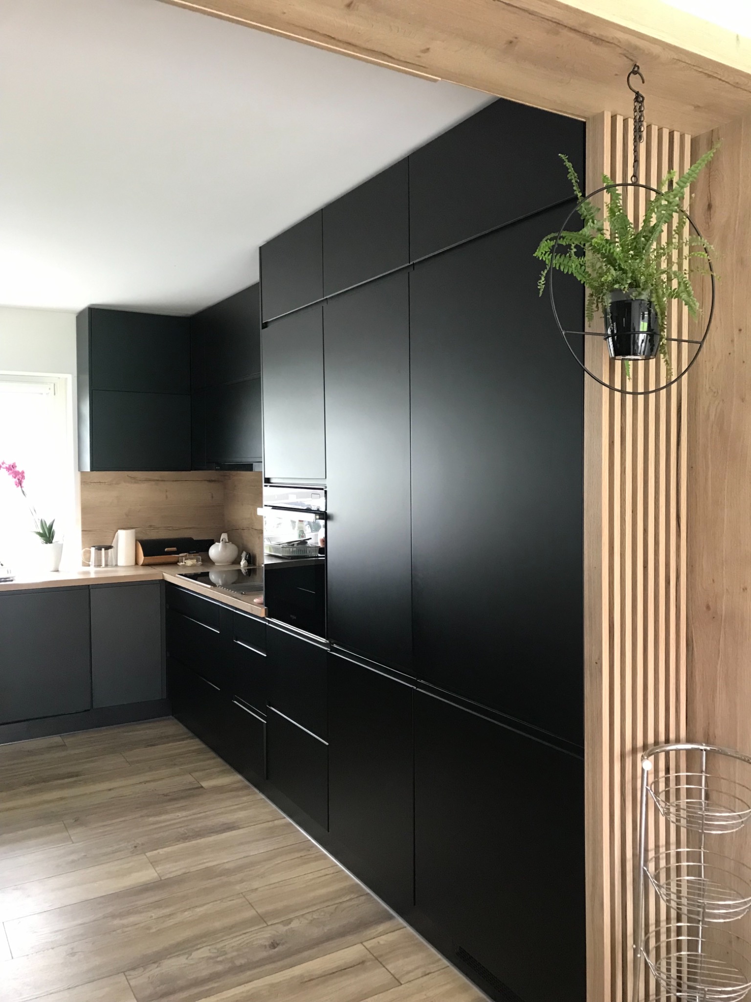 Nowoczesna kuchnia z czarnymi, matowymi frontami szafek, drewnianym blatem i podłogą imitującą drewno. Na ścianie drewniane lamele i wisząca roślina w metalowym kwietniku.