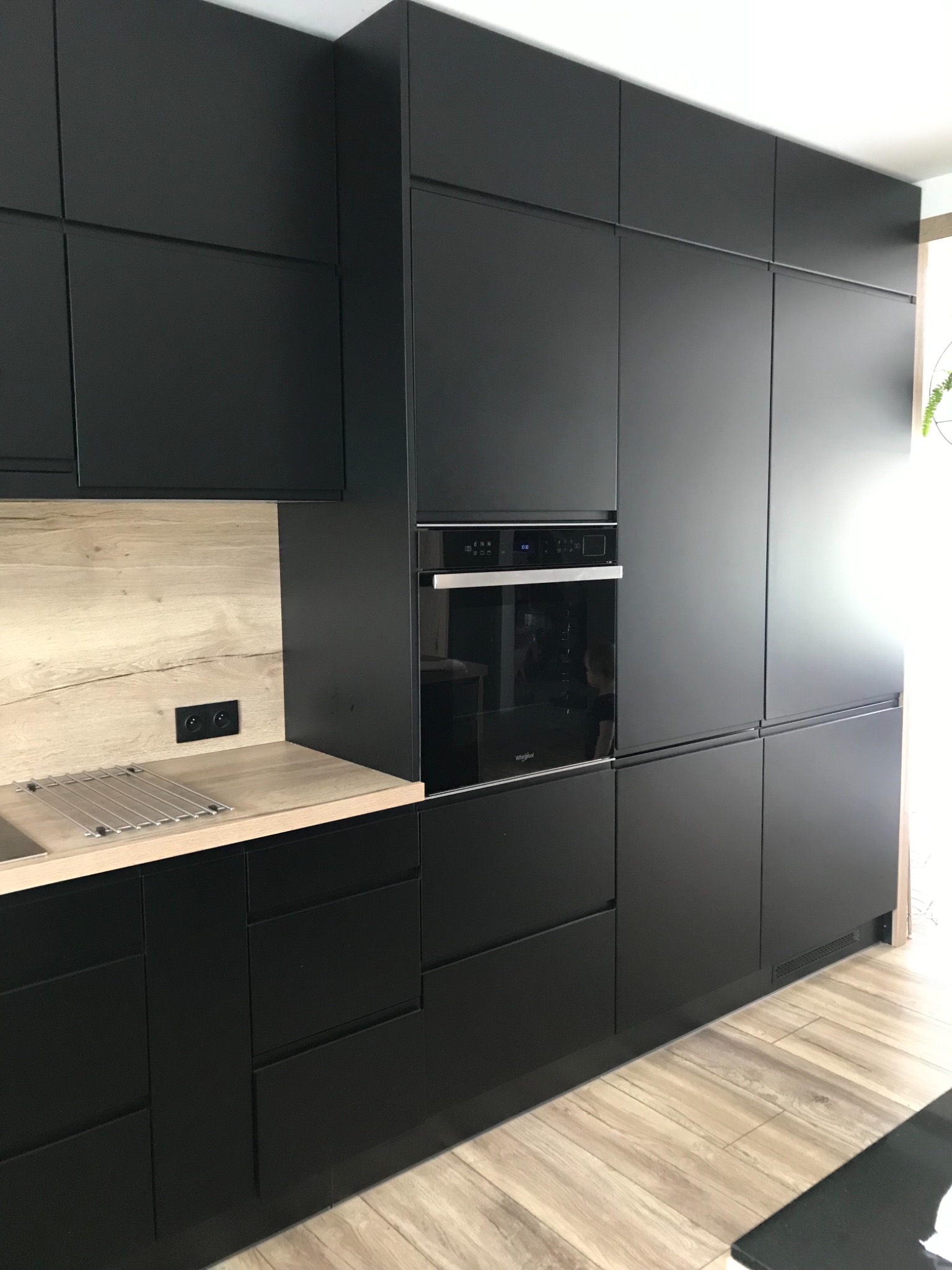 Część zabudowy kuchennej z matowymi, czarnymi frontami, drewnianym blatem i piekarnikiem, widoczna metalowa suszarka na naczynia.
