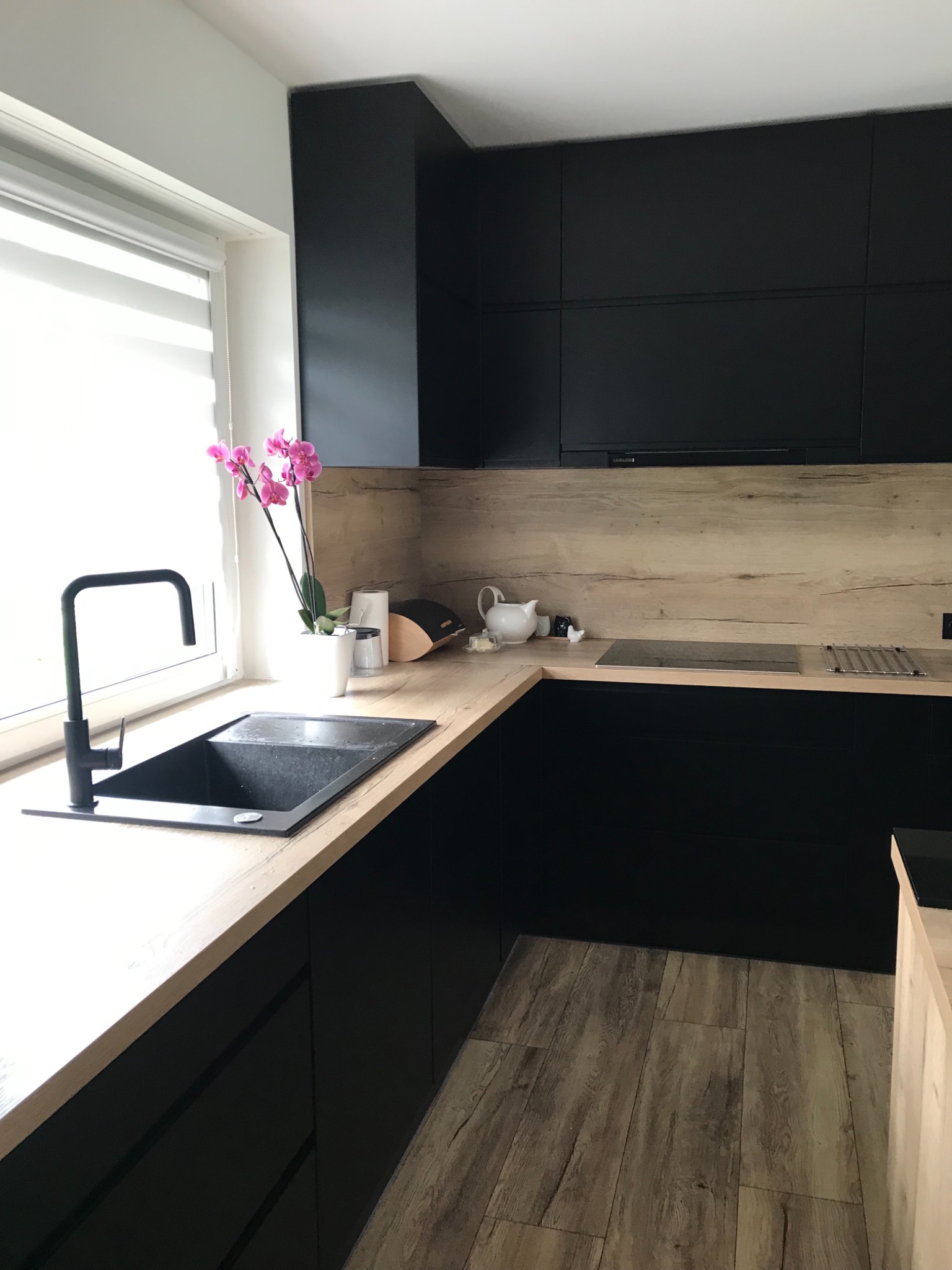 Nowoczesna kuchnia z czarnymi szafkami i blatem imitującym drewno, czarny zlew i bateria, z akcentem różowych kwiatów w doniczce przy oknie.