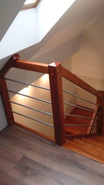 Drewniane schody z balustradą z poziomych metalowych prętów, oświetlone światłem wpadającym przez okno dachowe.