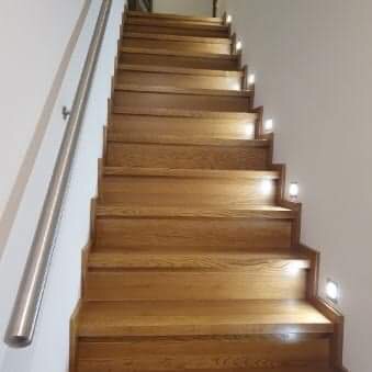 Schody drewniane z oświetleniem punktowym LED w ścianie, widok z dołu, poręcz ze stali nierdzewnej po lewej stronie.