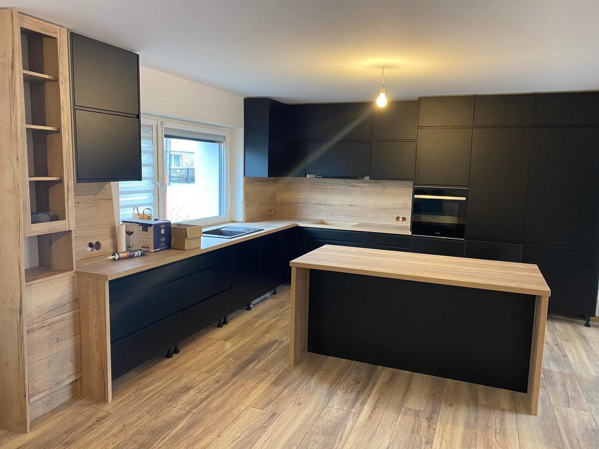 Nowoczesna kuchnia z czarnymi frontami i drewnianymi akcentami, wyspa kuchenna z blatem w kolorze drewna, parkiet imitujący drewno, minimalistyczne oświetlenie.