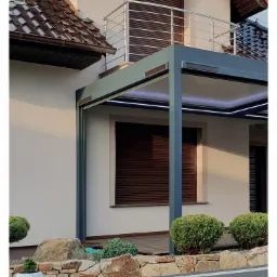 Nowoczesna pergola z szarymi słupami i oświetleniem LED, brązowe rolety zewnętrzne w oknach, kamienny murek z ozdobnymi krzewami w ogrodzie.