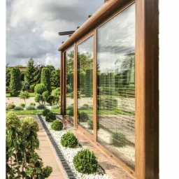 Przesuwne okna w kolorze brązu z widocznymi żaluzjami wewnętrznymi, otoczone zadbanym ogrodem z białymi kamykami i ozdobnymi krzewami.