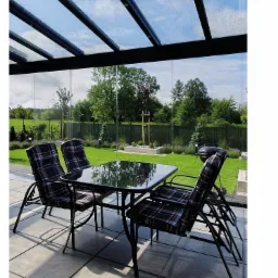 Zadaszony taras z przeszklonym dachem, widok na zadbany ogród, cztery krzesła z poduszkami w kratę i szklany stół.