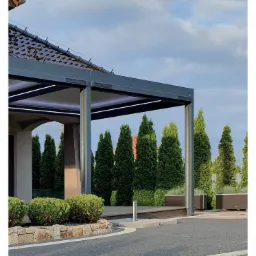 Nowoczesna pergola tarasowa z oświetleniem LED, widok z zewnątrz na tle iglaków i kostki brukowej.