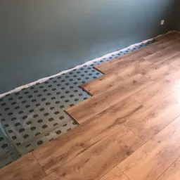 Fragment podłogi z drewnopodobnych paneli w trakcie montażu, widoczna podkładka izolacyjna z nadrukiem, ściana w odcieniu szarozielonym.