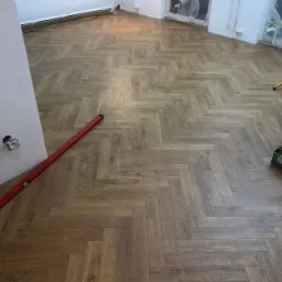 Ułożony parkiet w jodełkę francuską, widoczna poziomica przy ścianie, okno zabezpieczone folią.