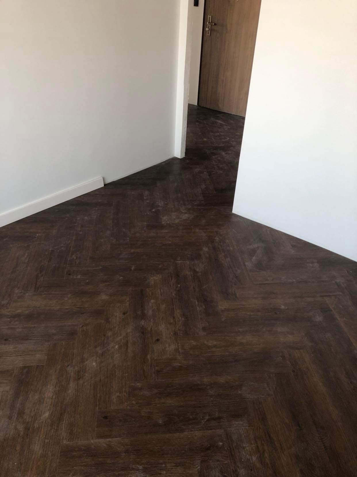 Ułożony parkiet w jodełkę w pokoju z białymi ścianami i drewnianymi drzwiami w tle.