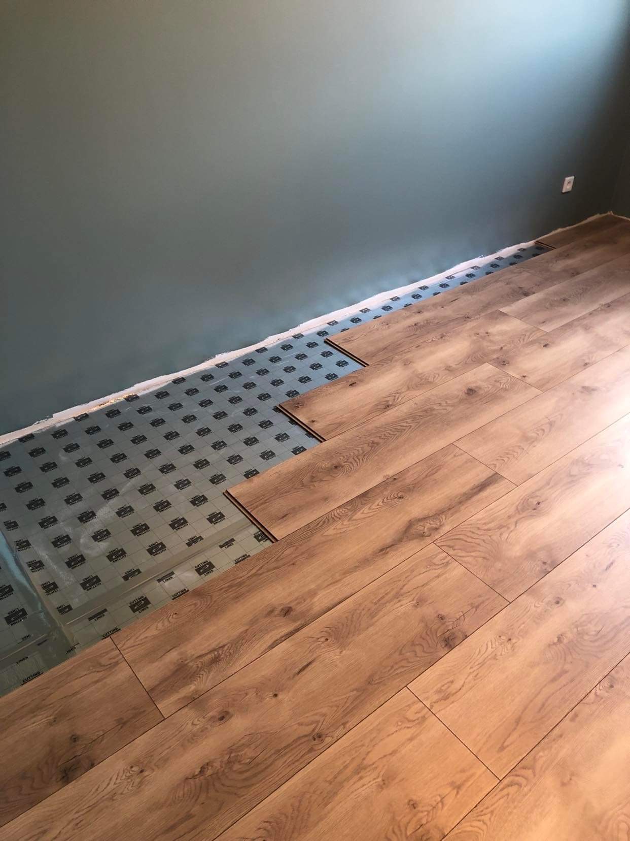 Fragment podłogi z drewnopodobnych paneli w trakcie montażu, widoczna podkładka izolacyjna z nadrukiem, ściana w odcieniu szarozielonym.