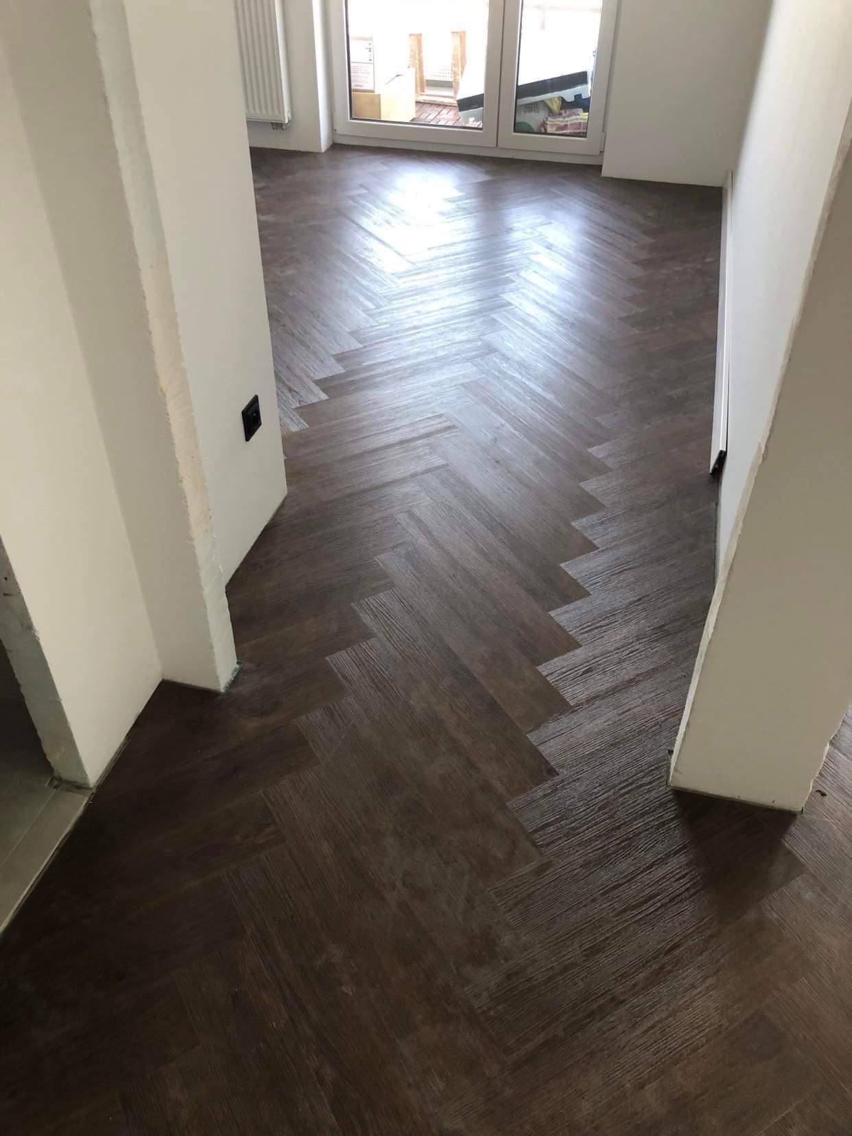 Ułożony parkiet w jodełkę w odcieniach brązu, widok z góry na fragment pomieszczenia z białymi framugami drzwi i widocznym fragmentem okna w tle.