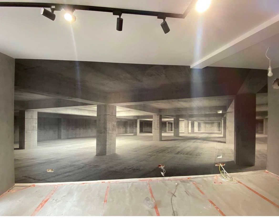 Ściana z fototapetą przedstawiającą surowy, betonowy parking podziemny; widoczne przewody elektryczne i lampy sufitowe.