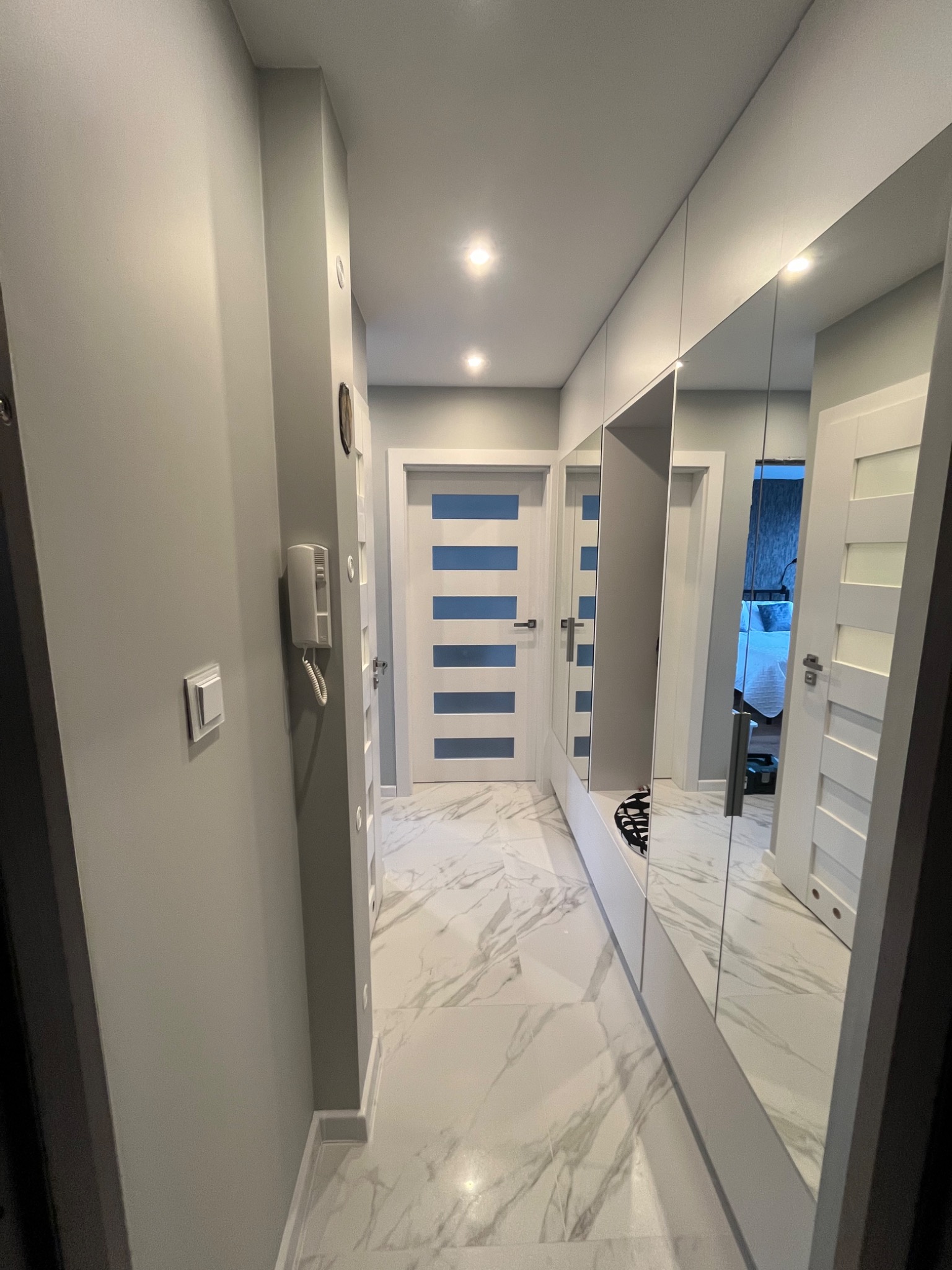 Wnętrze korytarza z szarą ścianą, marmurową podłogą, lustrzaną szafą wnękową i białymi drzwiami z niebieskimi wstawkami.