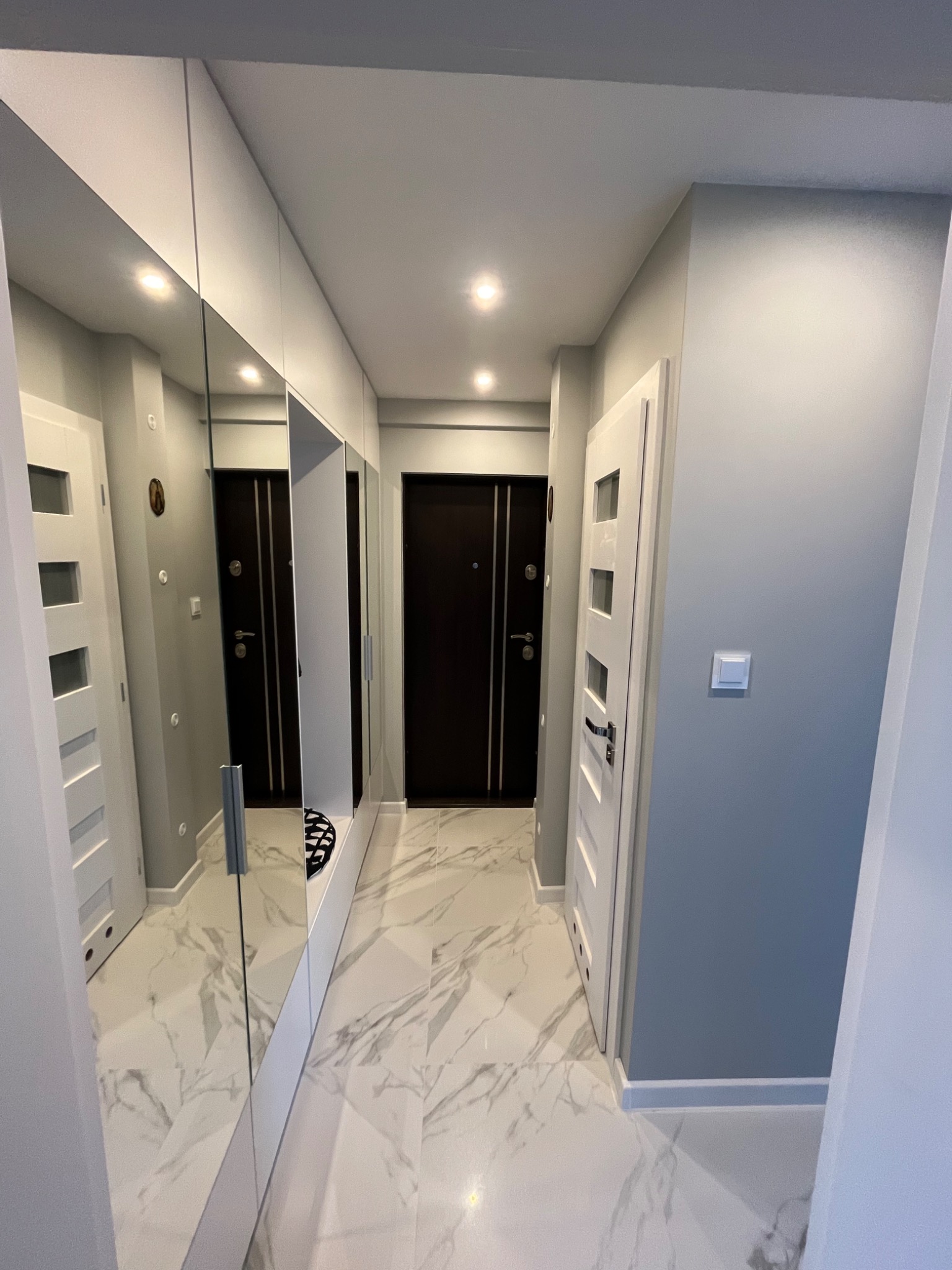 Nowoczesny przedpokój z szarymi ścianami, białymi drzwiami, lustrzaną szafą w zabudowie i marmurową podłogą z widocznym odbiciem czarnej wycieraczki w lustrze.