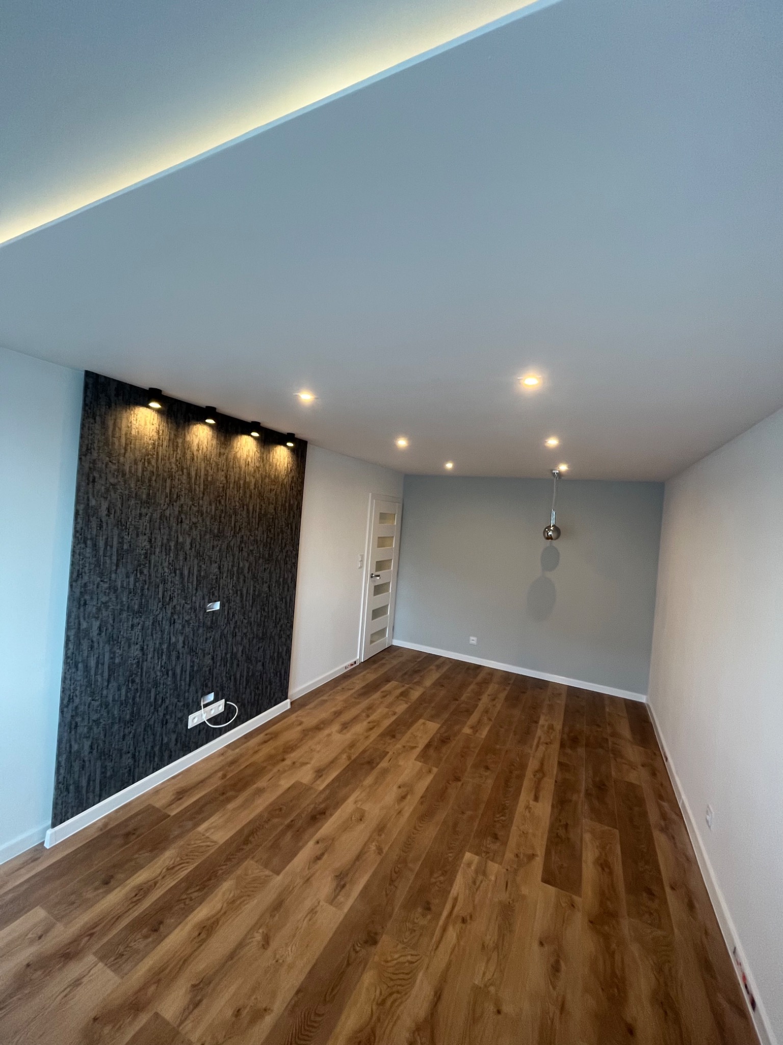 Nowoczesne wnętrze po remoncie: drewniana podłoga, ściana akcentowa z ciemną tapetą strukturalną oświetlona punktowo, jasne ściany, sufit z oświetleniem LED.