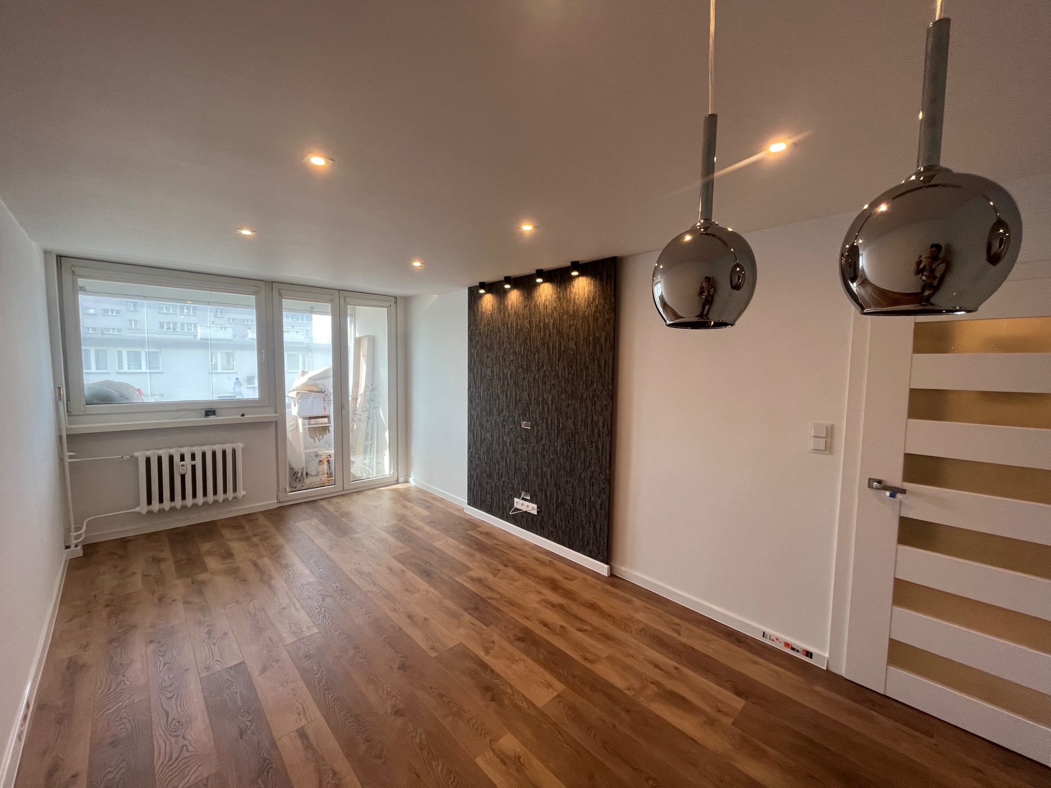 Jasne, przestronne wnętrze po remoncie z drewnianą podłogą, białymi ścianami, grzejnikiem pod oknem, dekoracyjną ścianą z oświetleniem punktowym i nowoczesnymi lampami wiszącymi.