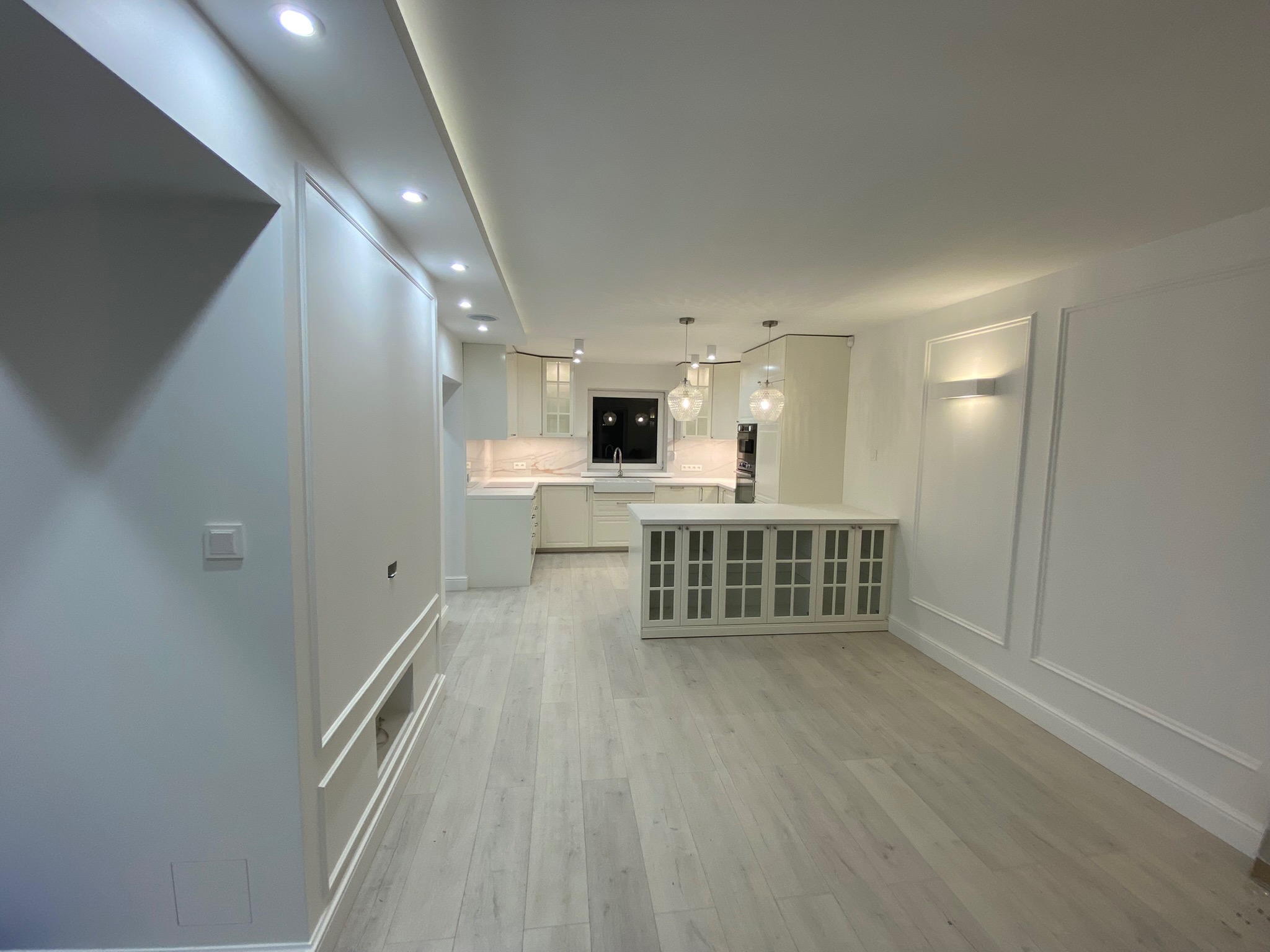 Jasne, przestronne wnętrze po remoncie: widok na kuchnię z wyspą, białe ściany z dekoracyjnymi listwami, drewniana podłoga, oświetlenie punktowe i wiszące lampy.