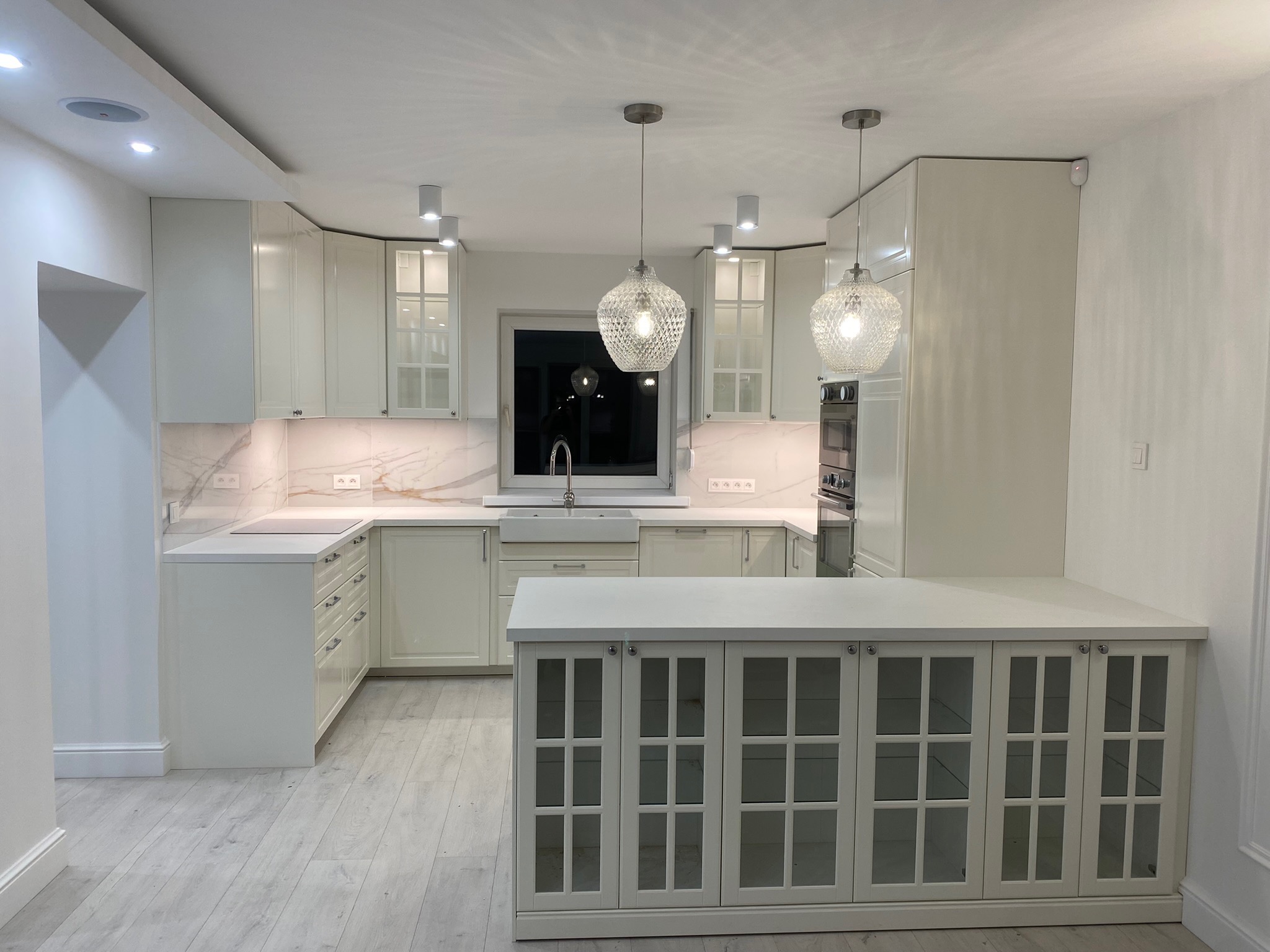 Elegancka, kremowa kuchnia z wyspą, oświetlona wiszącymi lampami z kryształkami, z marmurowym blatem i frontami szafek z przeszklonymi witrynami.