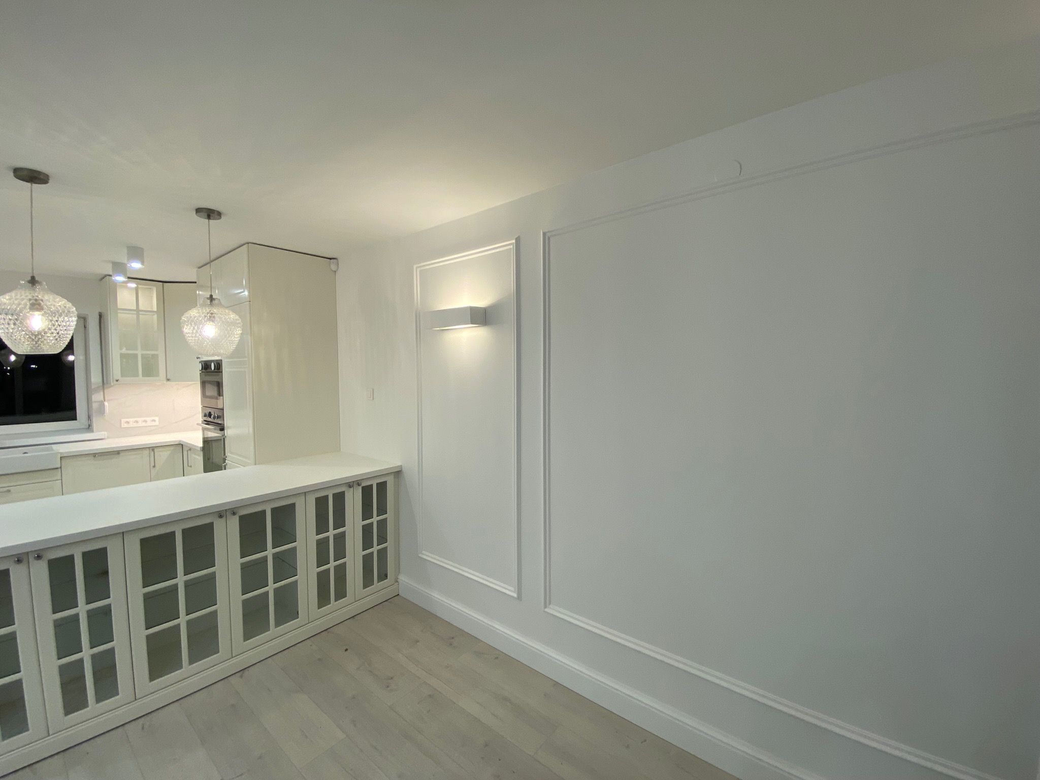 Jasne wnętrze po remoncie: widok na ścianę z dekoracyjnymi listwami i oświetleniem, oraz fragment kuchni z przeszklonymi szafkami i wiszącymi lampami.