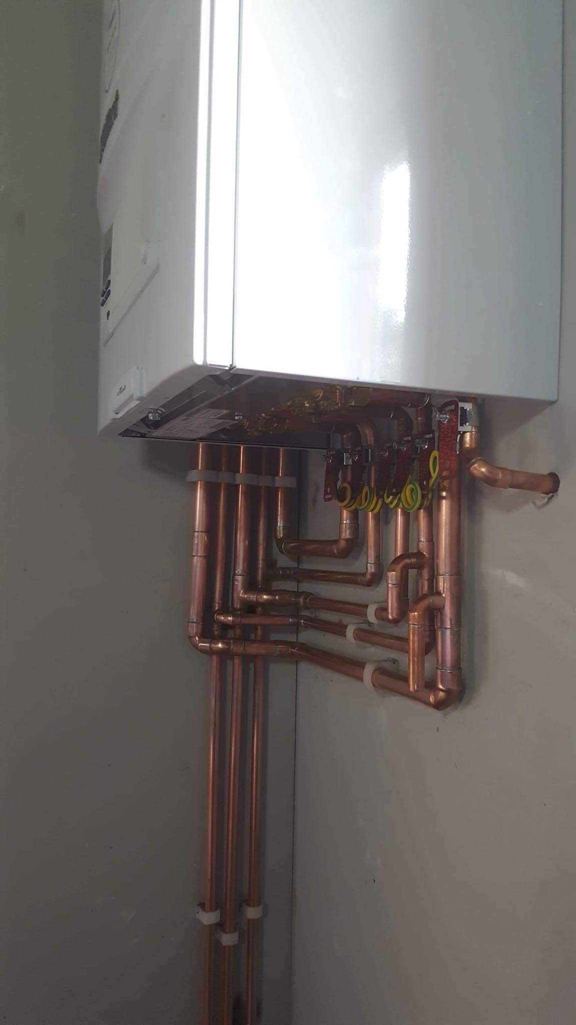 Biały piec centralnego ogrzewania zamontowany na ścianie, z widocznymi miedzianymi rurami i przewodami elektrycznymi, zabezpieczonymi białymi uchwytami.