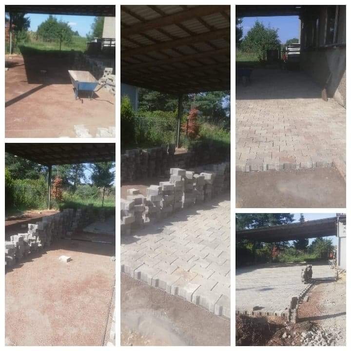 Układanie kostki brukowej w zadaszonej przestrzeni: widok na częściowo ułożoną nawierzchnię z szarej kostki, stosy kostki przygotowanej do ułożenia oraz taczkę w tle.