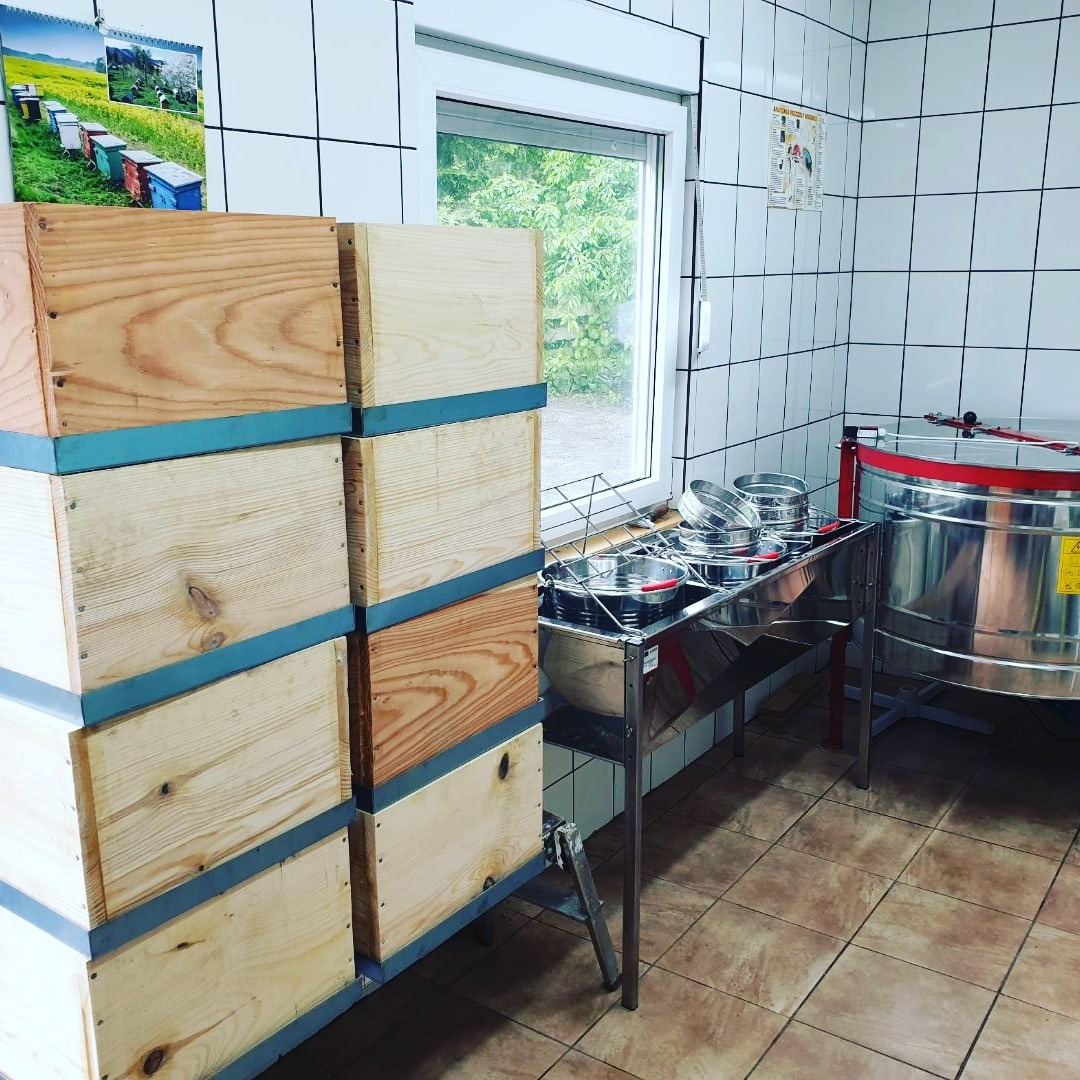Stos drewnianych uli z niebieskimi paskami, obok stalowy stół z sitami i miodarka w pomieszczeniu z kafelkami.