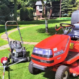 Czerwony traktorek do trawy marki Kubota HST, kosiarka spalinowa NAC i podkaszarka spalinowa leżące na trawniku w ogrodzie z domkiem dla dzieci i huśtawką w tle.