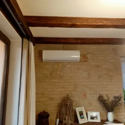 Klimatyzator ścienny zamontowany w salonie z ceglaną ścianą i drewnianymi belkami stropowymi, widok z dołu.