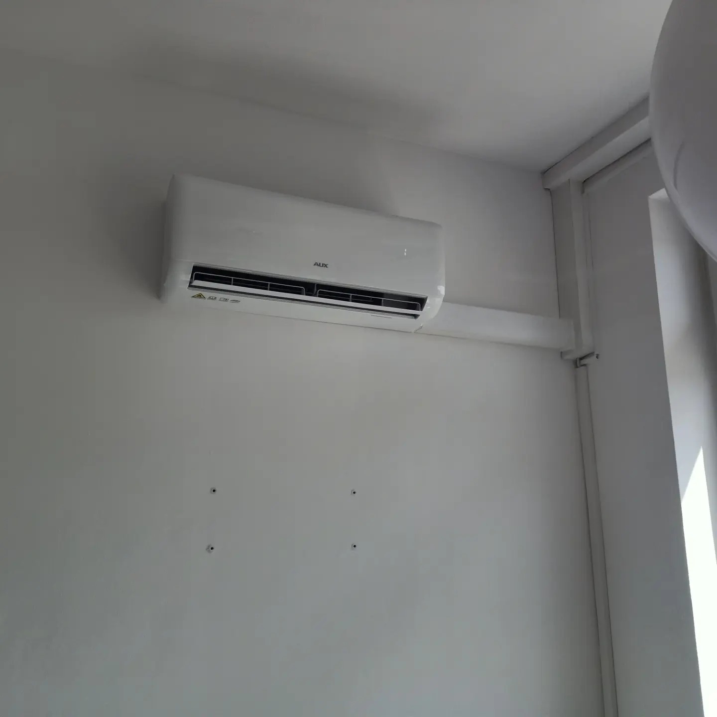 Biały klimatyzator marki AUX zamontowany na białej ścianie w pomieszczeniu. Widoczne otwory po wcześniejszych mocowaniach na ścianie.
