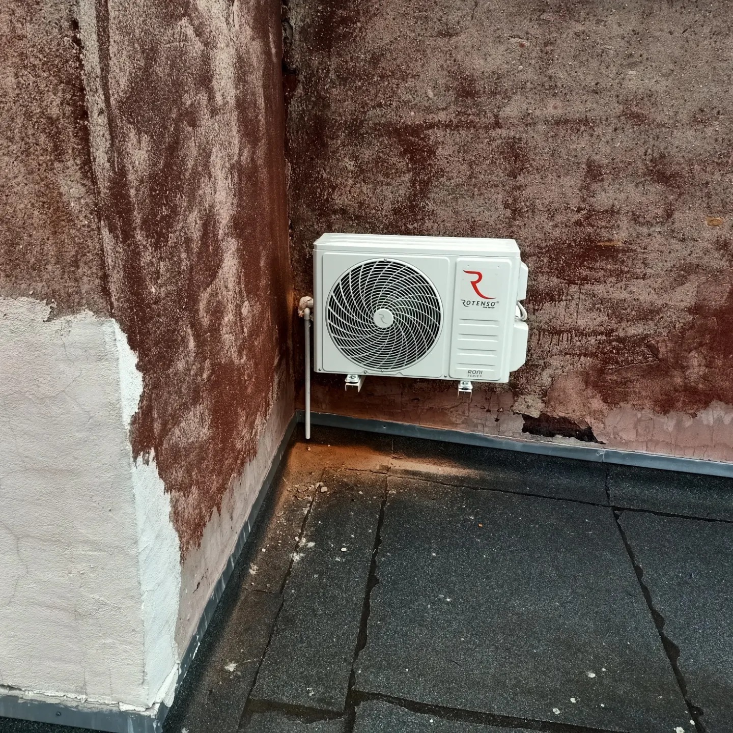 Biała jednostka zewnętrzna klimatyzacji Rotenso zamontowana na zewnątrz budynku z brudnymi ścianami i gumową podłogą.