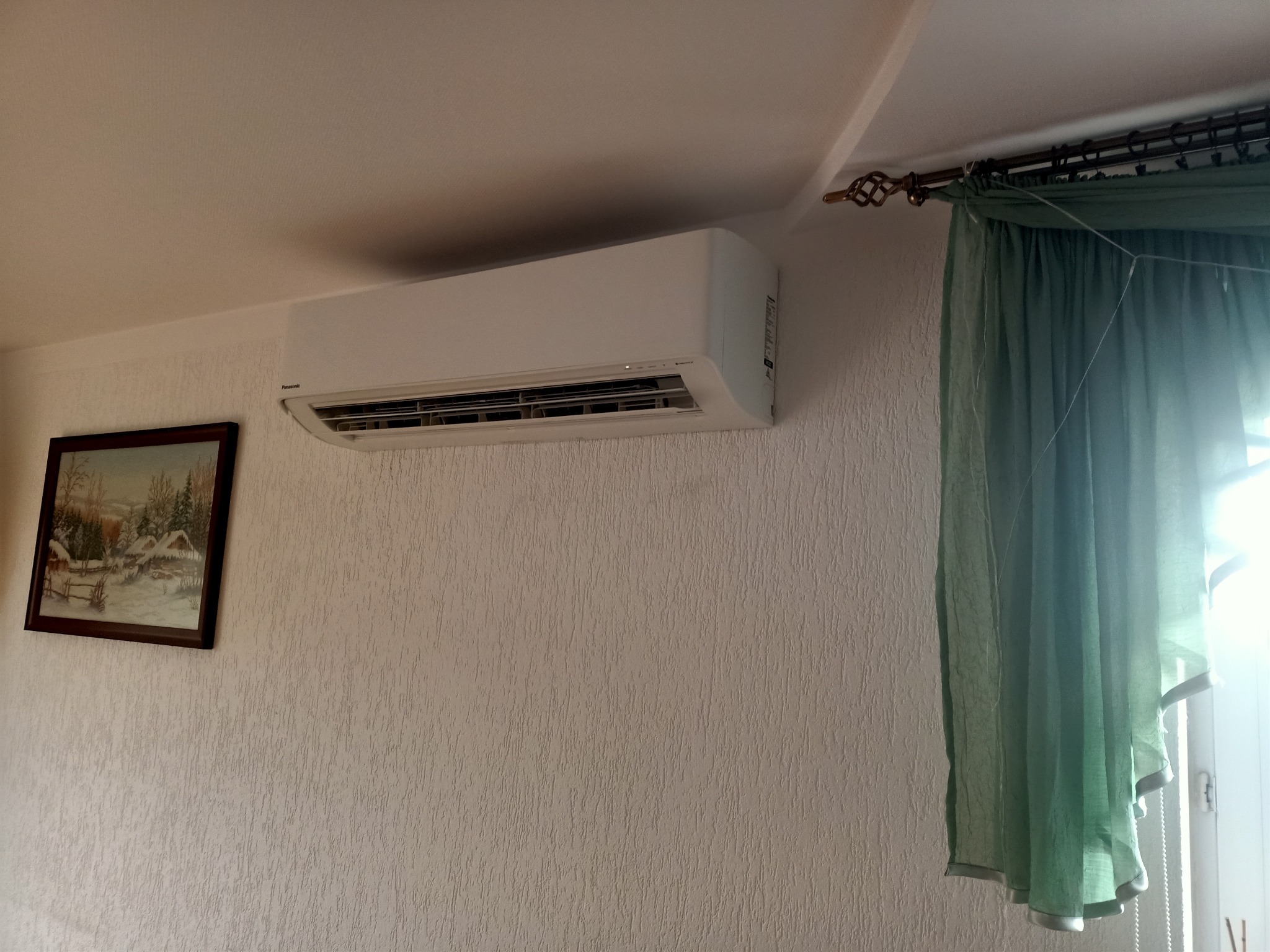 Biały klimatyzator ścienny marki Panasonic zamontowany w pokoju z teksturowaną tapetą, obok obrazu zimowego krajobrazu i zasłoniętego okna.
