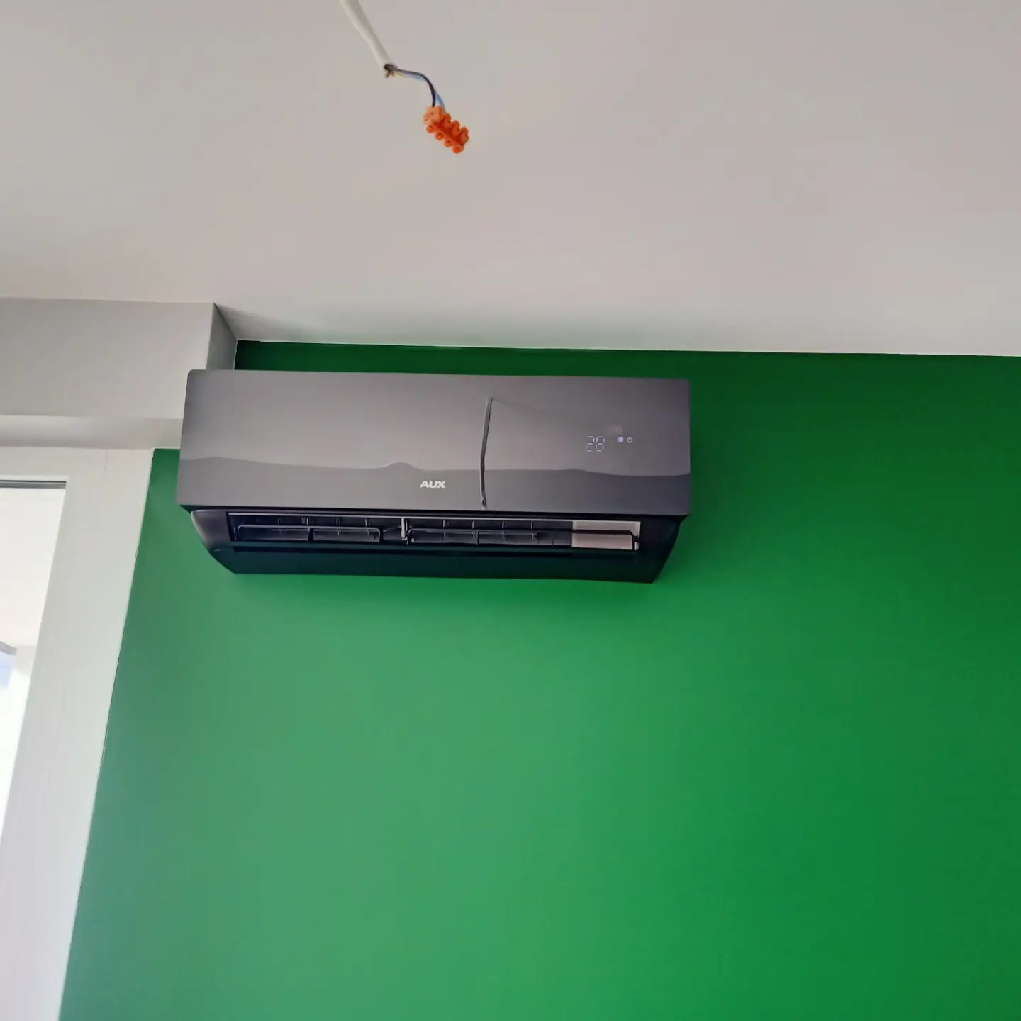 Szary klimatyzator AUX zamontowany na jaskrawo zielonej ścianie, z widocznymi przewodami elektrycznymi wystającymi z sufitu.