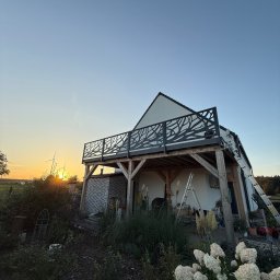 Usługi Stolarskie Lofthomeland Jakub Korzeniowski - Dom z tarasem i balustradą o wzorze drzewa na tle zachodzącego słońca. Widoczne drabiny sugerują prace wykończeniowe. W tle turbiny wiatrowe.