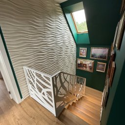 Usługi Stolarskie Lofthomeland Jakub Korzeniowski - Nowoczesne schody z jasnego drewna z białą, ażurową balustradą, oświetlone od góry oknem dachowym. Ściana z dekoracyjną fakturą i galeria obrazów w ciemnozielonym wnętrzu.