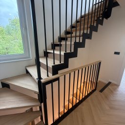 Usługi Stolarskie Lofthomeland Jakub Korzeniowski - Nowoczesne schody zabiegowe z jasnymi, szerokimi stopniami i czarną, geometryczną konstrukcją metalową oraz pionową balustradą. Naturalne światło z okna, podłoga z jasnego drewna w jodełkę.