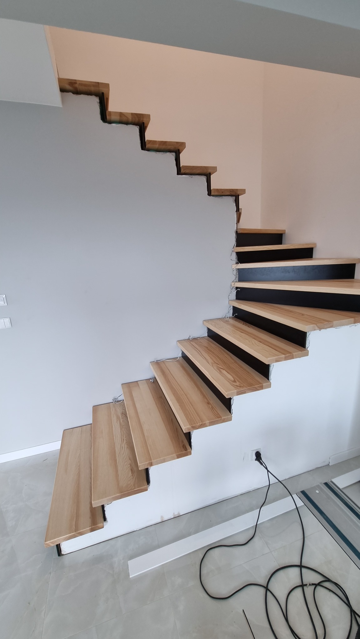 Montaż drewnianych schodów z czarnymi podstopnicami, widoczne niedokończone wykończenie ściany i przewody elektryczne.