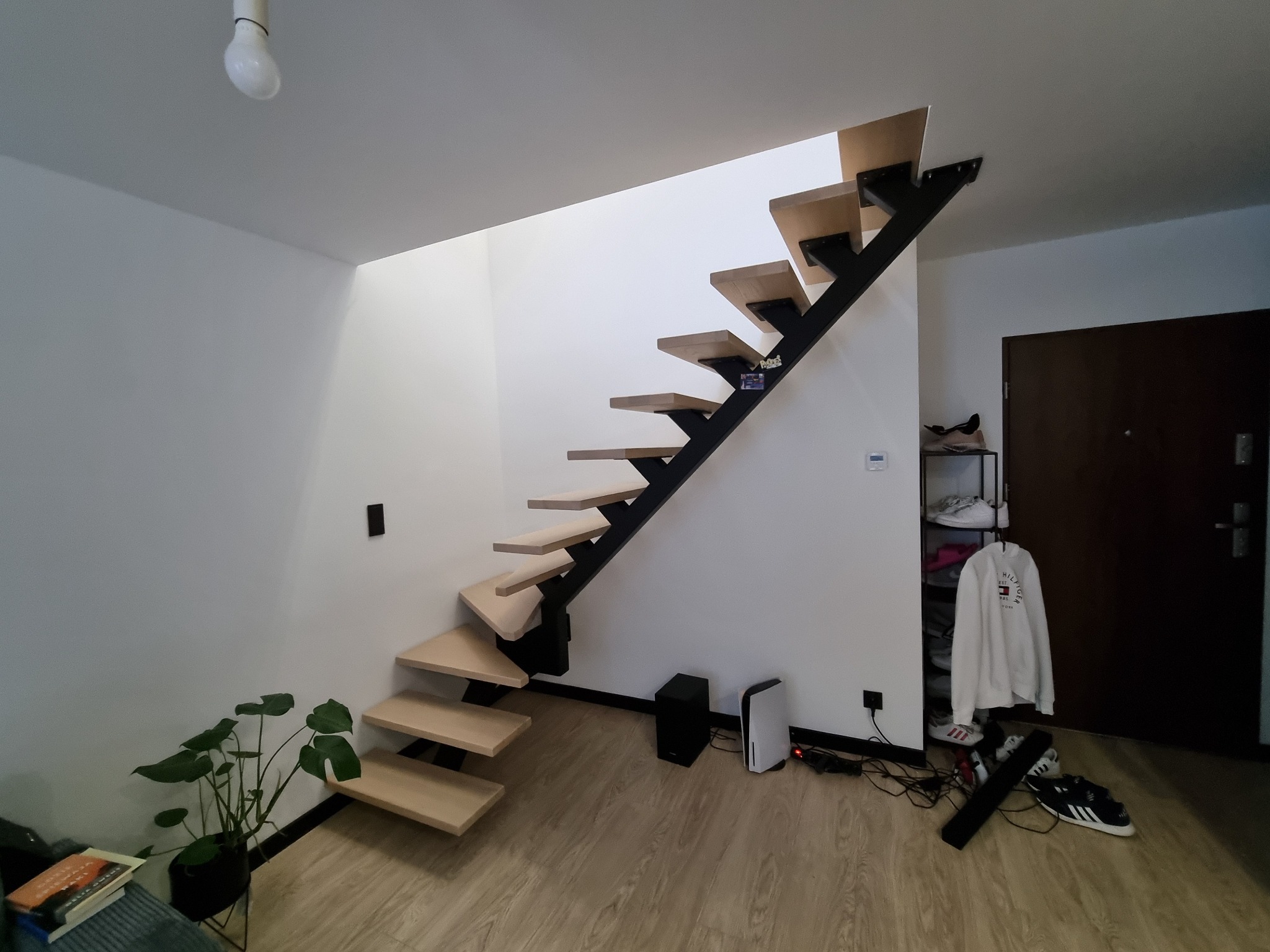 Nowoczesne schody z drewnianymi stopniami i czarną metalową konstrukcją w jasnym wnętrzu z rośliną doniczkową i sprzętem elektronicznym.