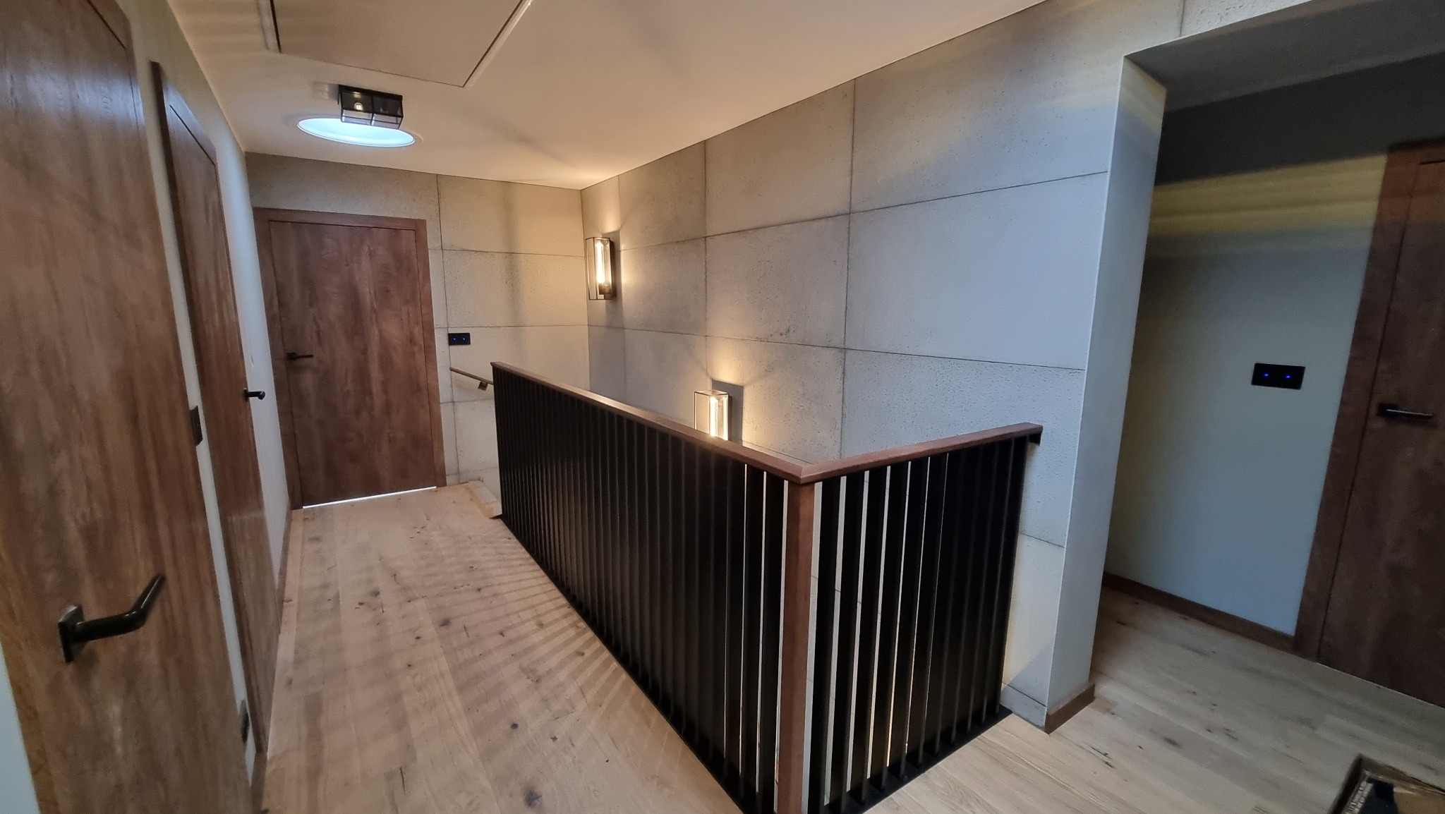 Nowoczesna balustrada schodowa z ciemnego drewna na jasnej podłodze, oświetlona kinkietami na teksturowanej ścianie, widok z góry schodów.