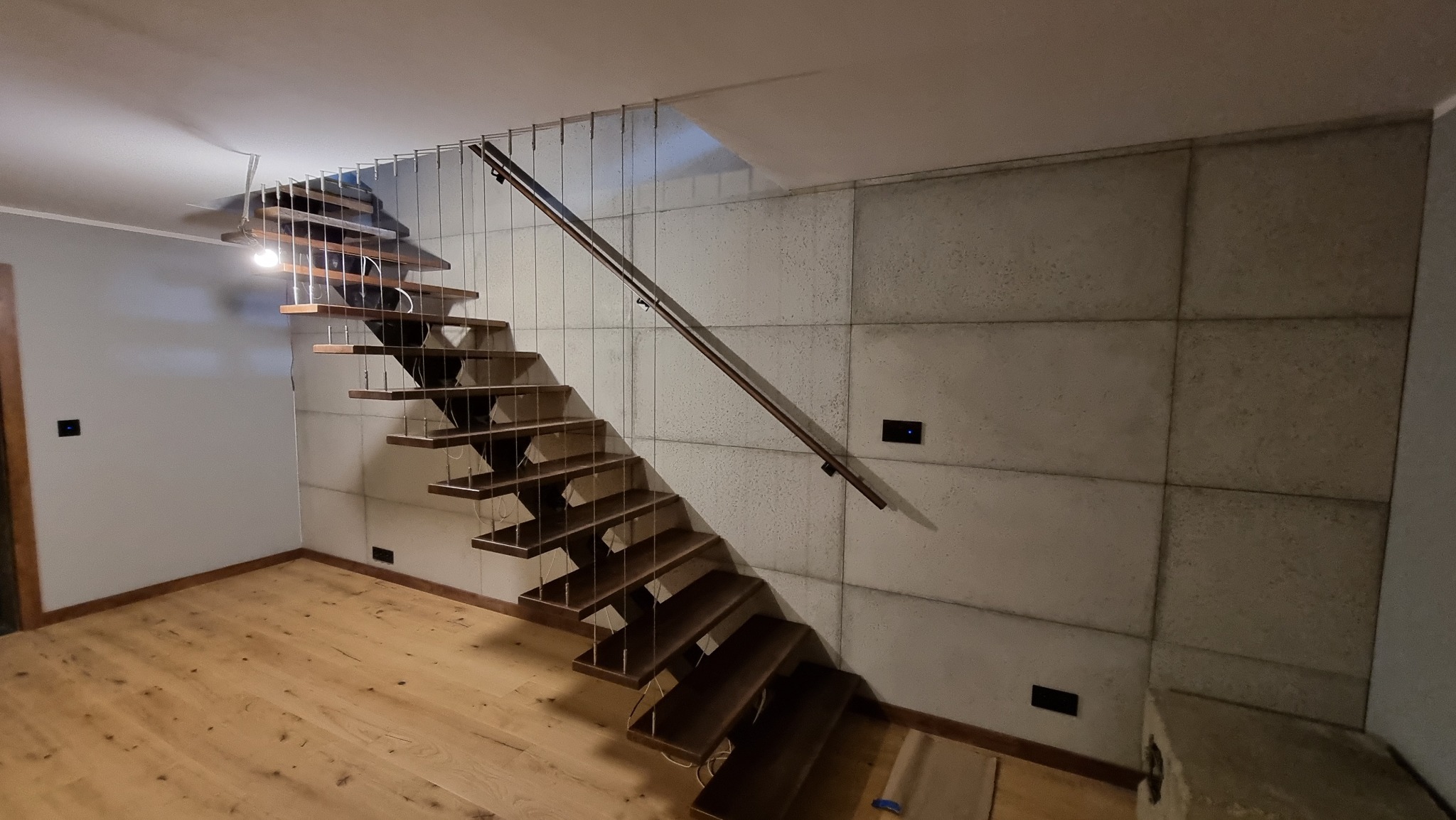 Nowoczesne schody z drewnianymi stopniami zawieszonymi na stalowych linkach, z pojedynczą poręczą zamocowaną do ściany wyłożonej kwadratowymi panelami.