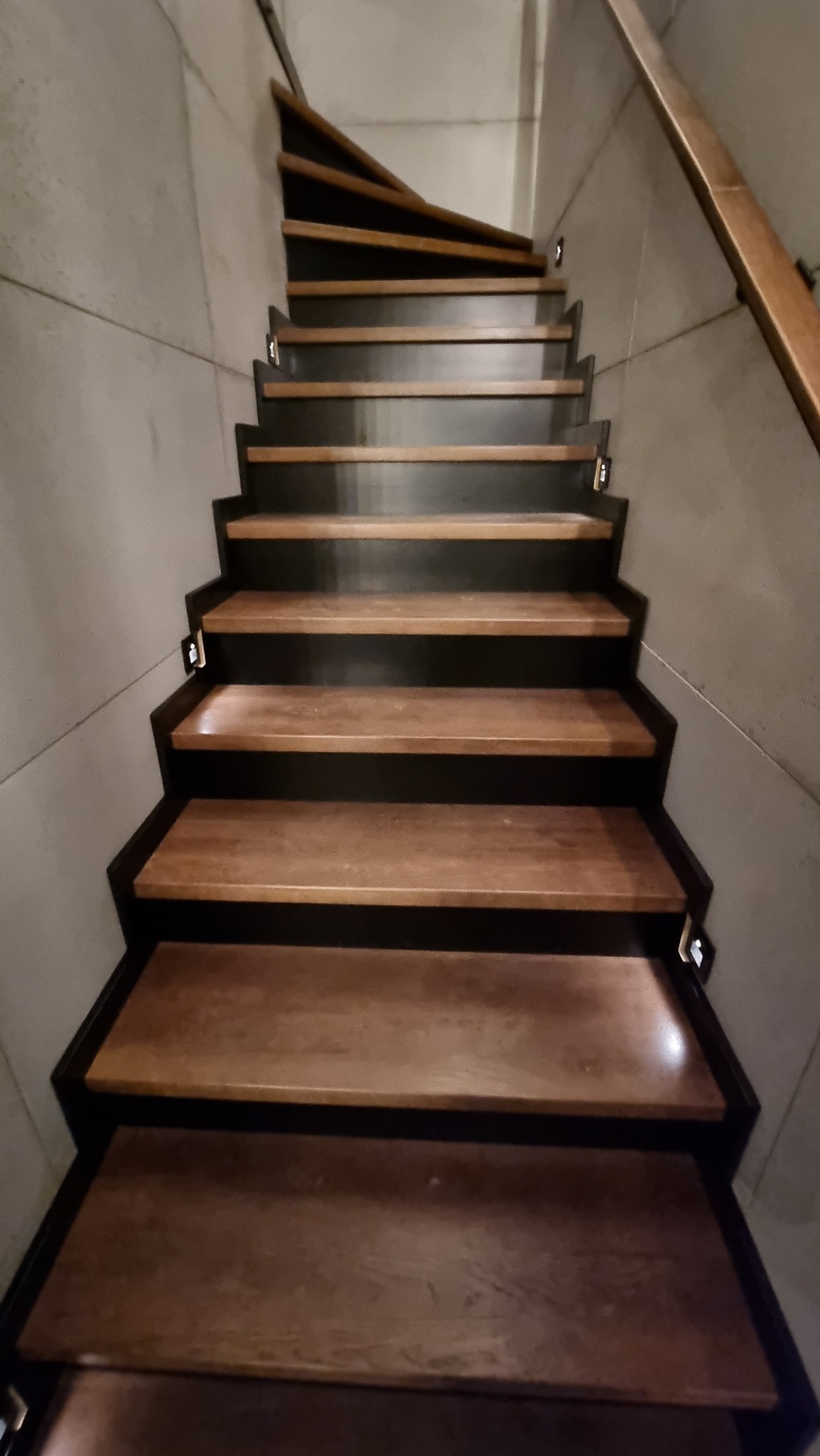 Wewnętrzne schody z drewnianymi stopniami i czarnymi podstopnicami, oświetlone małymi lampkami LED przy ścianie, prowadzące w górę po skręcie.