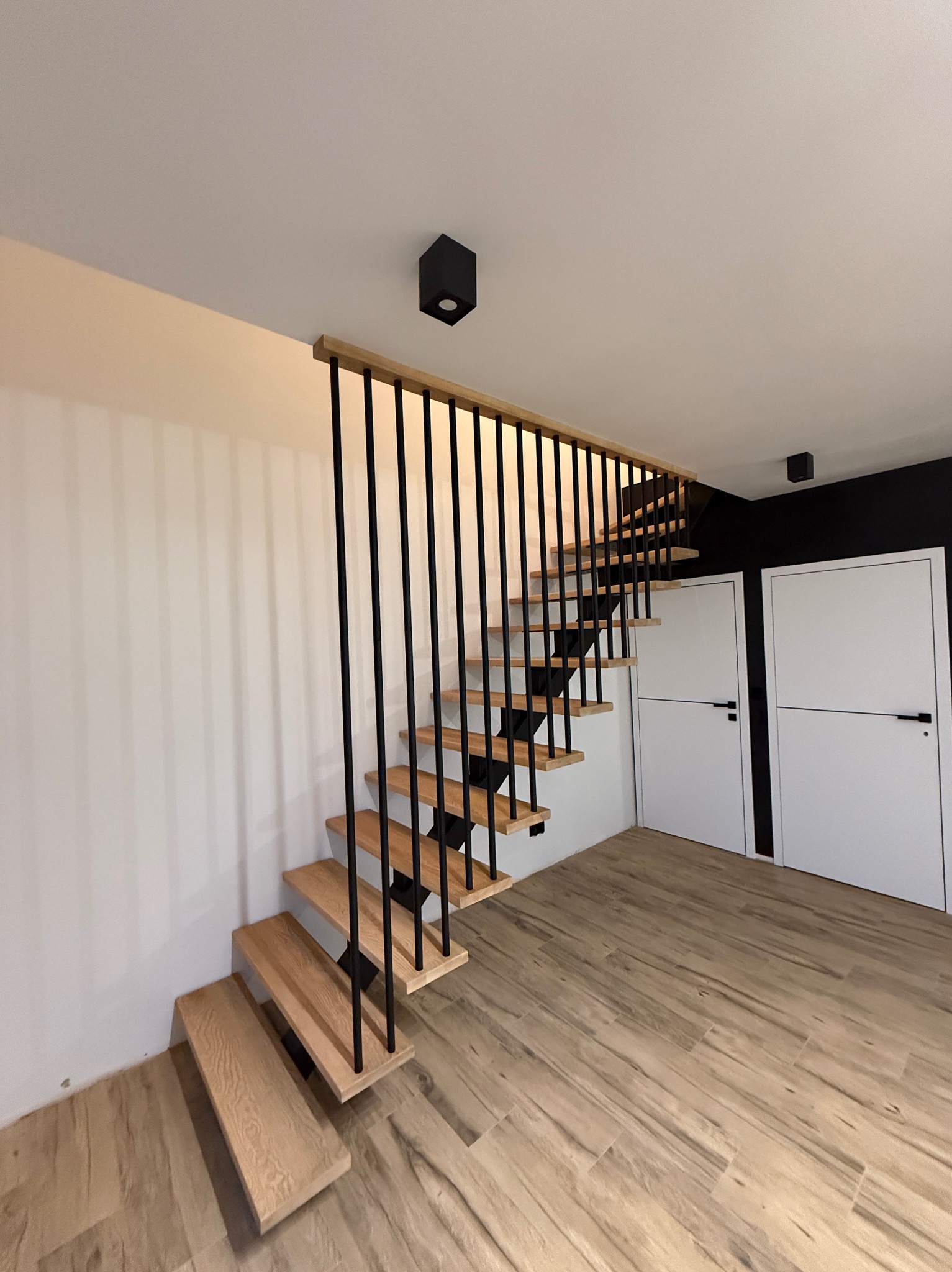 Nowoczesne schody drewniane z czarnymi, metalowymi balustradami w jasnym wnętrzu z drewnianą podłogą i dwoma białymi drzwiami w tle.