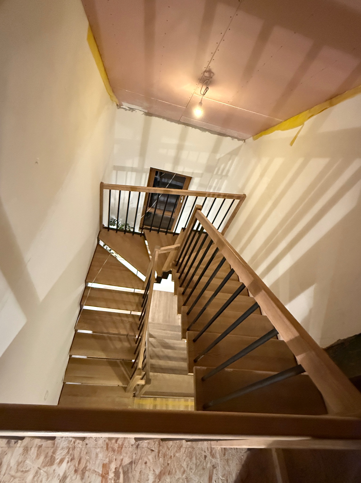 Wysokie ujęcie schodów z drewnianymi stopniami i czarnymi metalowymi balustradami w trakcie montażu, widoczne niedokończone wykończenie ścian i sufitu, surowy stan budowy.