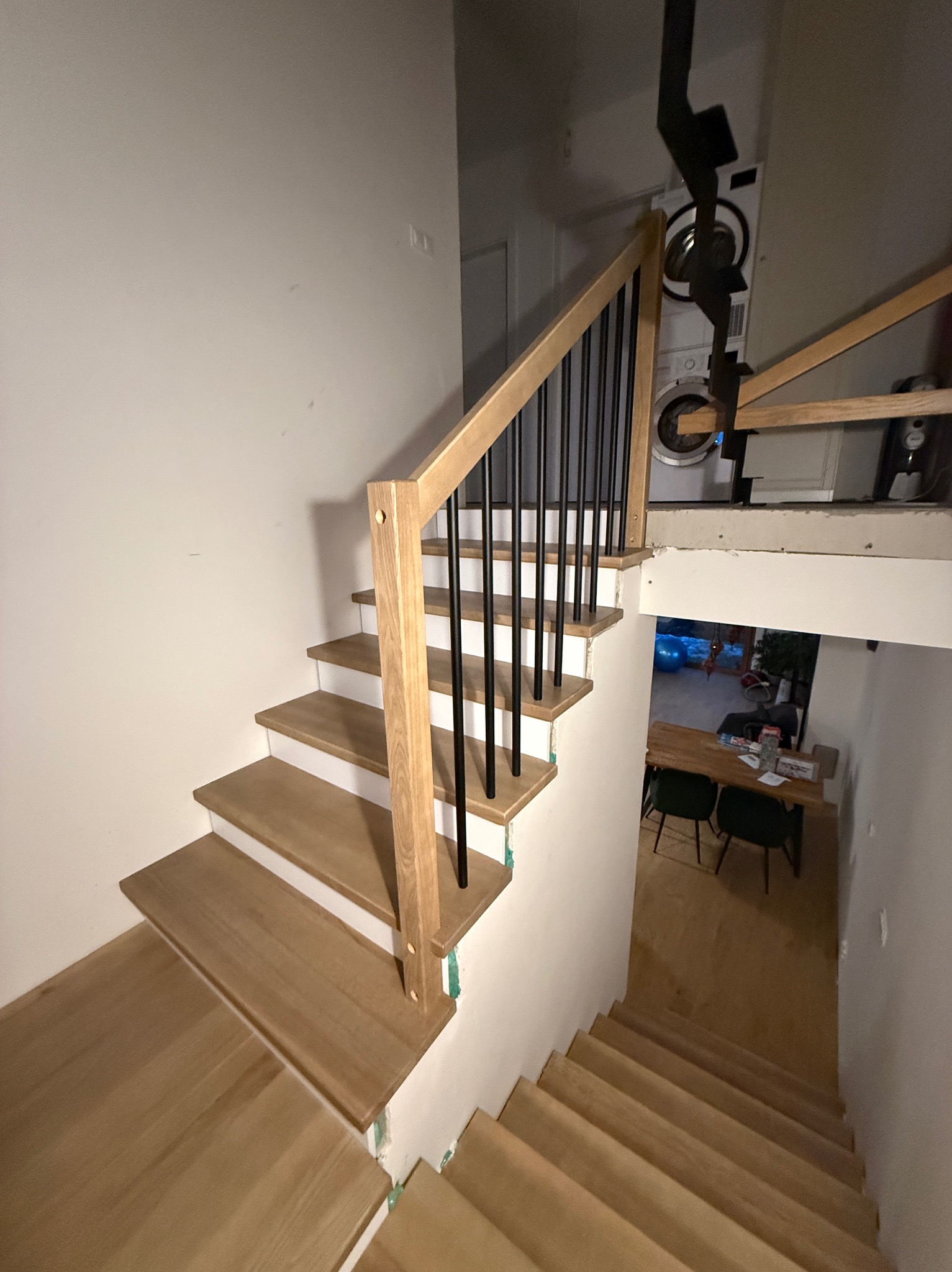 Nowe schody z jasnego drewna z czarnymi, metalowymi balustradami w trakcie montażu, widoczne zabezpieczenia na ścianach i pralka w tle.