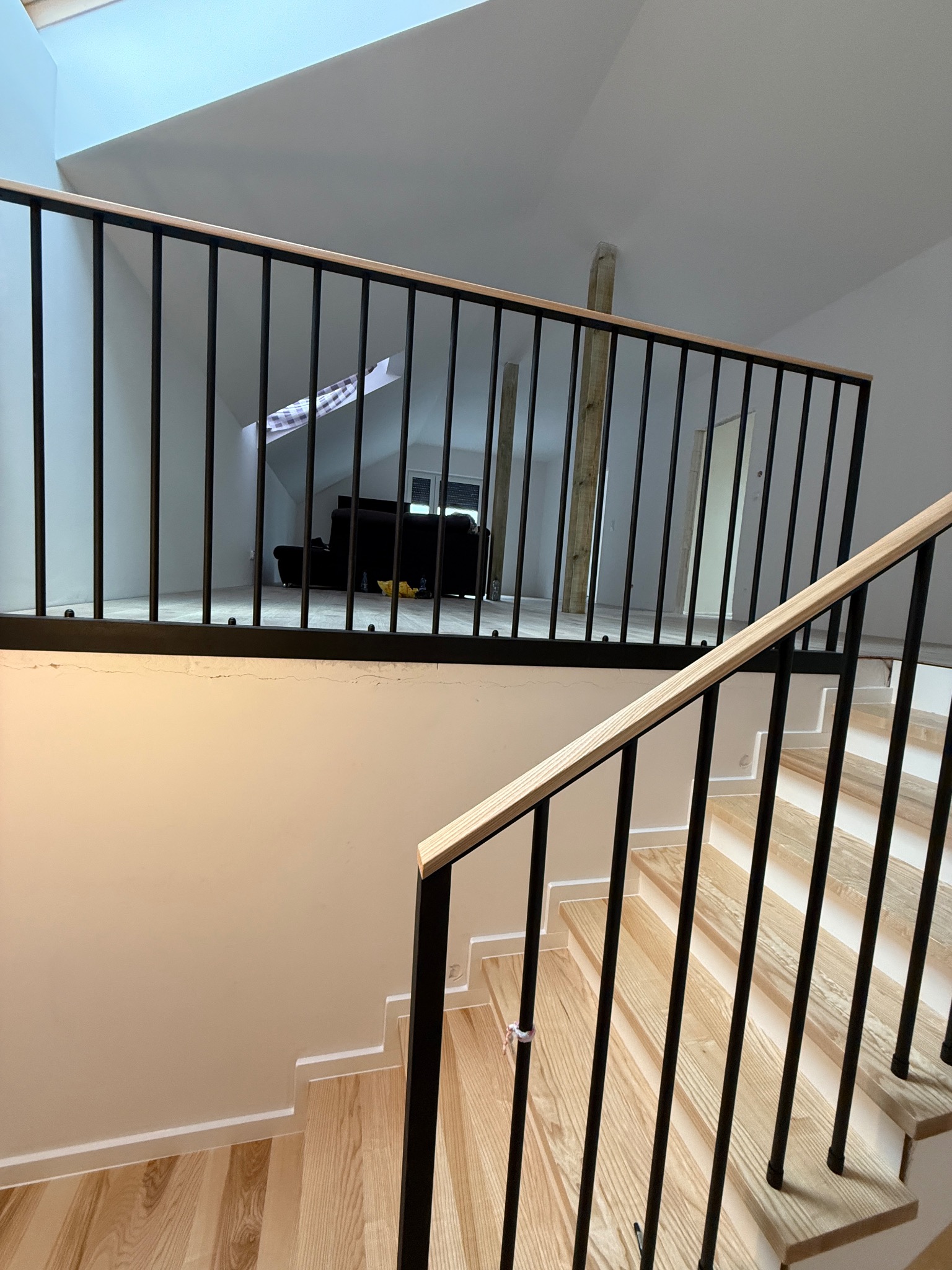 Jasne schody z drewnianymi stopniami i czarną, metalową balustradą z drewnianym poręczem. Widok z góry na dół, w tle jasne pomieszczenie na poddaszu.